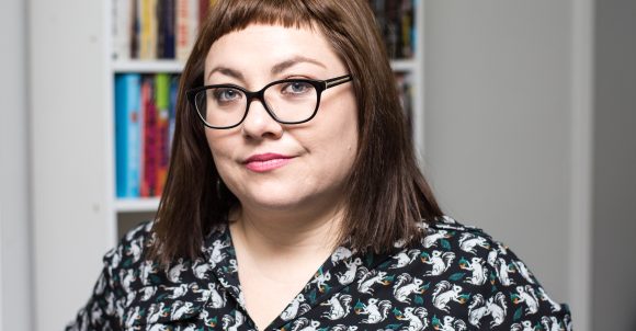 Isabel Quintero headshot. Isabel has straight, shoulder-length auburn hair with bangs and wears glasses and a black and white button-up shirt in front of bookshelves.