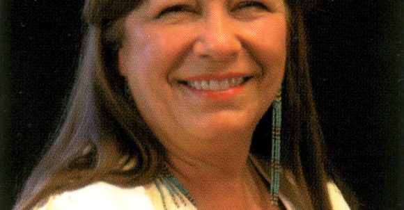 Jeanne Rorex Bridges headshot. Jeanne has long, brown hair with bangs, long earrings, and a white fringe top over a black shirt.