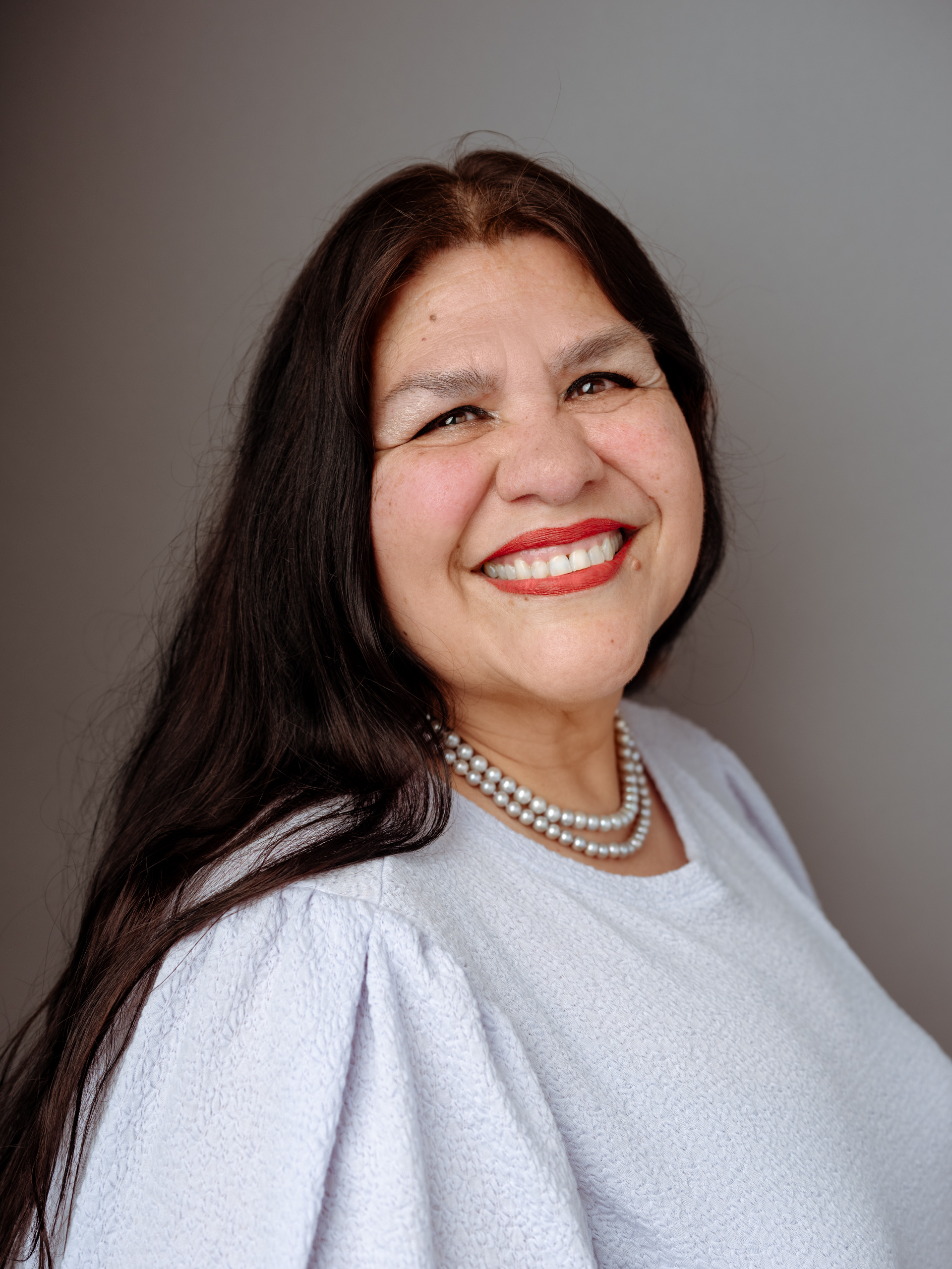 Guadalupe García McCall wearing a white blouse, pearl necklace, red lipstick and smiling with her shoulders turned toward the right