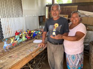 Avelino Pérez holds and stands next to his carvings. To his left is wife, Martina Morgado. Both stand and smile at the camera.