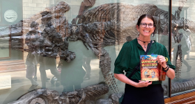 Cindy Jenson-Elliott holds up a copy of The Doomsday Detectives and stands In front of Mary Anning's Ichtheosaur fossil in the London Natural History Museum.