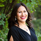 Photo of Dezi Lynn, an indigenous woman with shoulder-length, brown hair wearing a black top with a necklace and earrings standing in front of trees