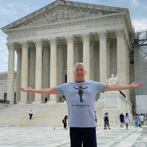 Author Michael Genhart stands in front of the US Supreme Court Building with his arms stretched wide and a smile on his face. He wears a gray t-shirt that has a silhouette of Edie Windsor on it. She wears a rainbow scarf. Below the silhouette is a quote attributed to her that says, “Don’t Postpone Joy!”
